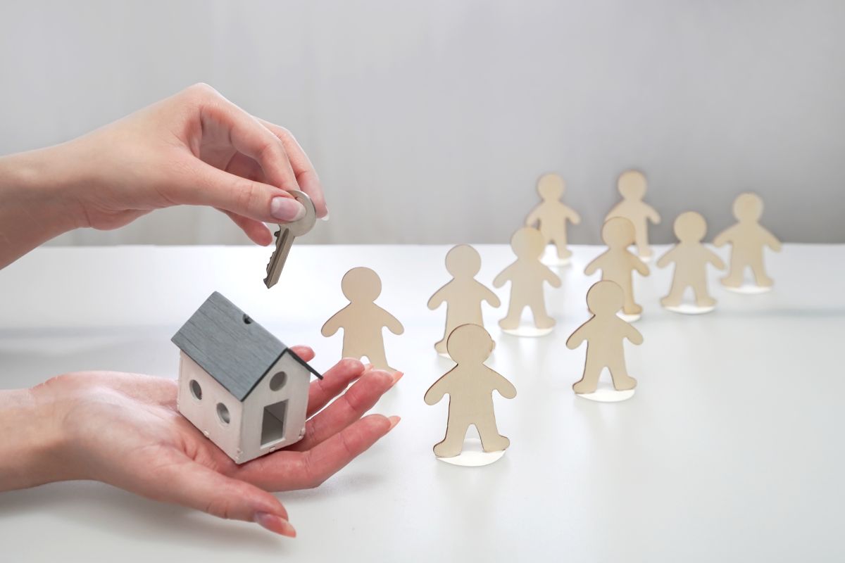 Main tenant une maquette de maison et une clé, l'offrant à des personnages en carton 
